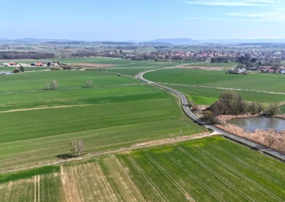Luftaufnahme: grüne Felder, kurvenreiche Straße, Teich, Bäume, Dorf, blauer Himmel.