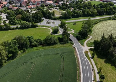 Eine asphaltierte Straße schlängelt sich durch grüne Felder zu rot gedeckten Häusern, der Himmel ist bewölkt.