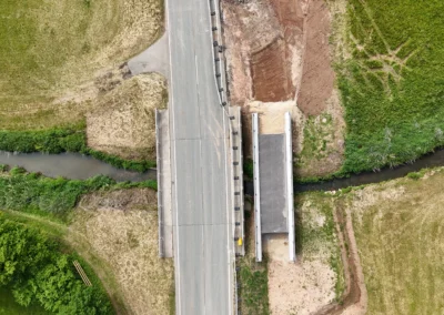 Luftaufnahme: Straßenbrücke, Bach, neuer Straßenabschnitt, Grasflächen in der Nähe.